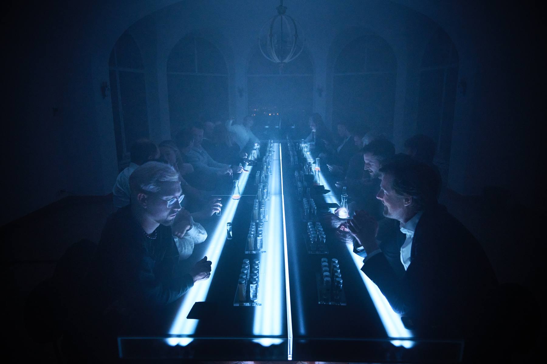 Guests seated along a long illuminated acrylic table in a completely dark room during Tabula Rasa